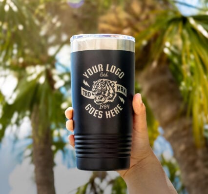 Hand Holding Custom Metal Engraved Tumbler That Reads "Your Logo Goes Here" The Hand Holding The Custom Tumbler Is In Front Of a Background of Palm Trees and A Blue Sky With White Clouds